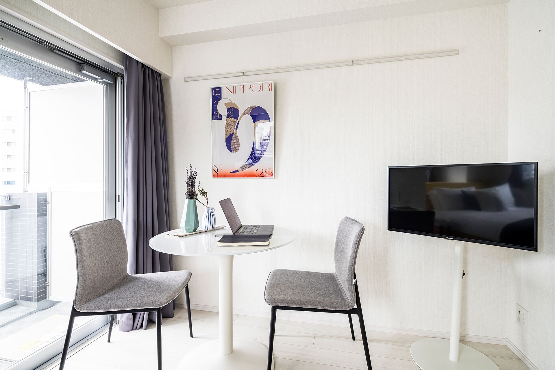Ein modernes Hotelzimmer mit großem Fenster, einem an der Wand montierten Flachbildfernseher, zwei grauen Stühlen um einen kleinen weißen Tisch und einem dekorativen Gegenstand über dem Fernseher.