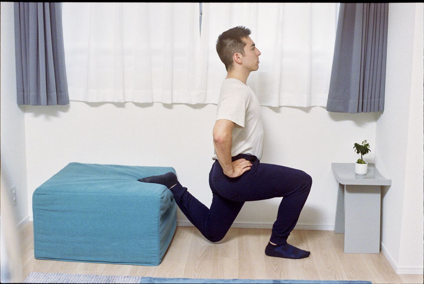A person performing a stretching exercise in a room with a blue ottoman, a small white table with a plant, and a window with curtains.