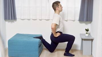 A person performing a stretching exercise in a room with a blue ottoman, a small white table with a plant, and a window with curtains.
