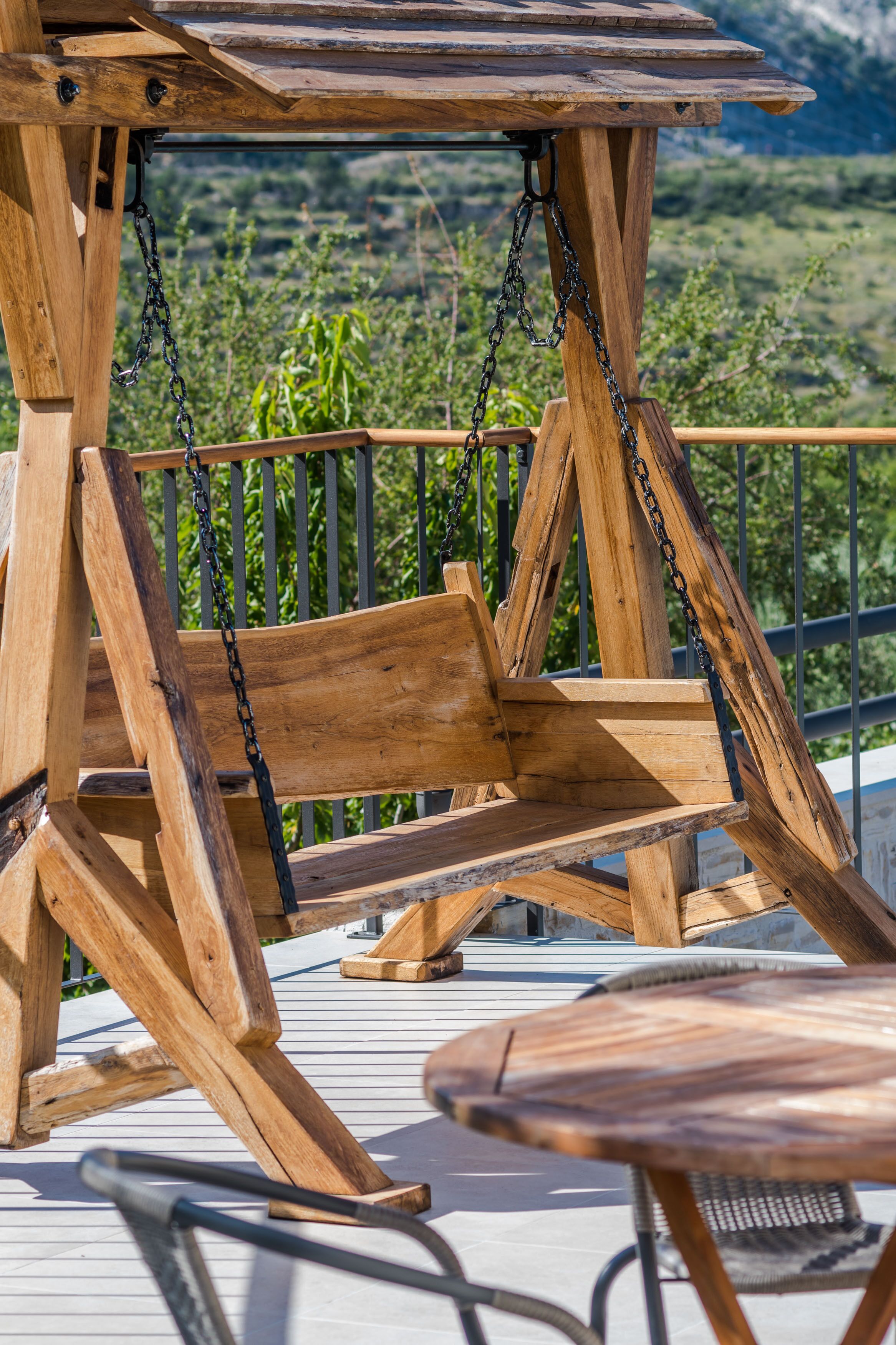 Una sedia a dondolo in legno con struttura metallica, fissata a una struttura in legno dotata di ringhiera metallica, con vista su un paesaggio verde.