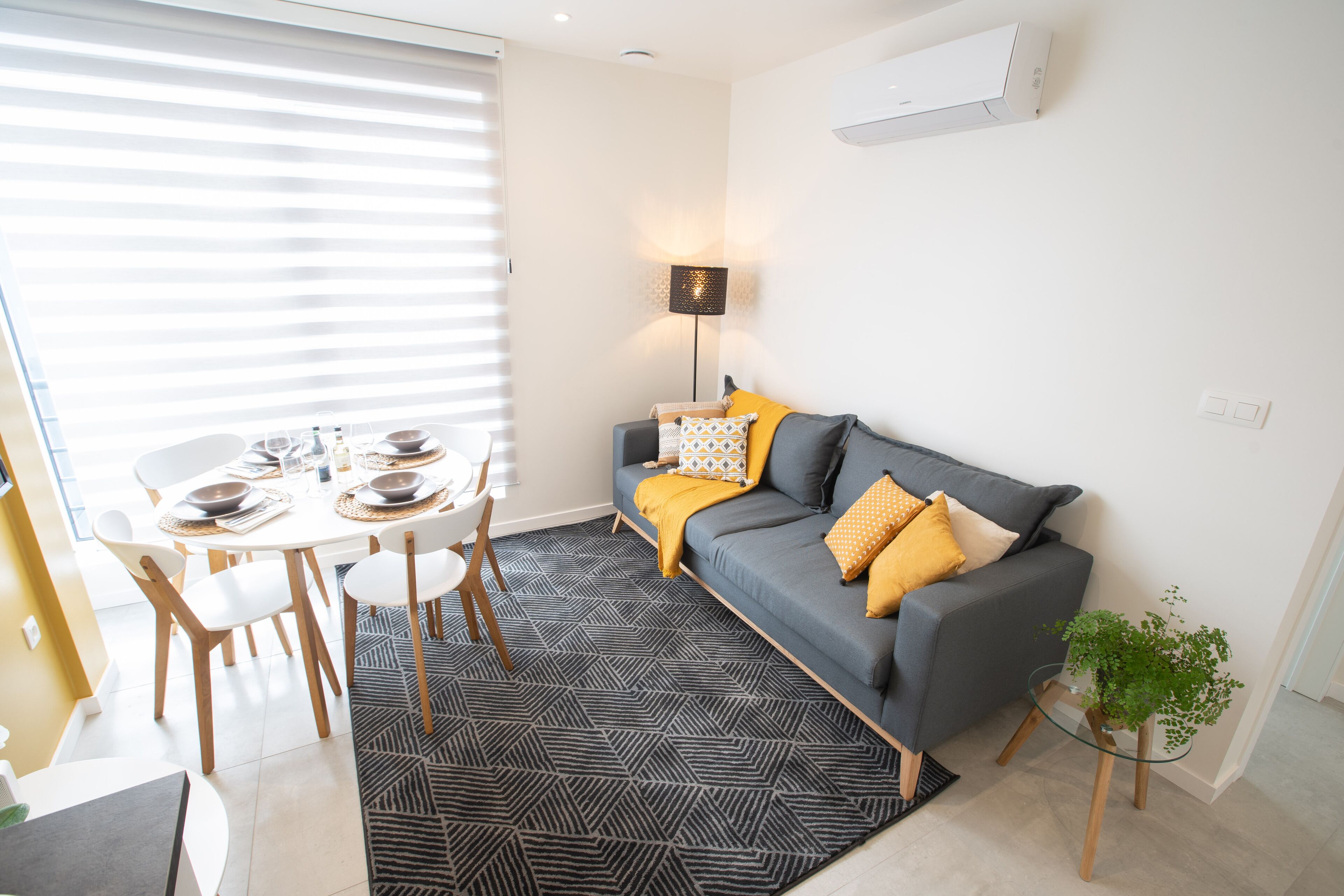A modern living room with a grey sofa, yellow cushions, a glass coffee table, and a dining area with a white table and chairs.