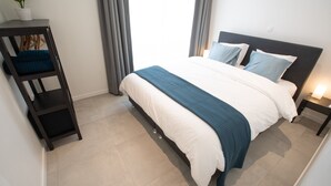 A modern bedroom with a bed, a nightstand, a dark wooden shelf with a plant, and a window with curtains. - Studio Brussels Airport (Zaventem)