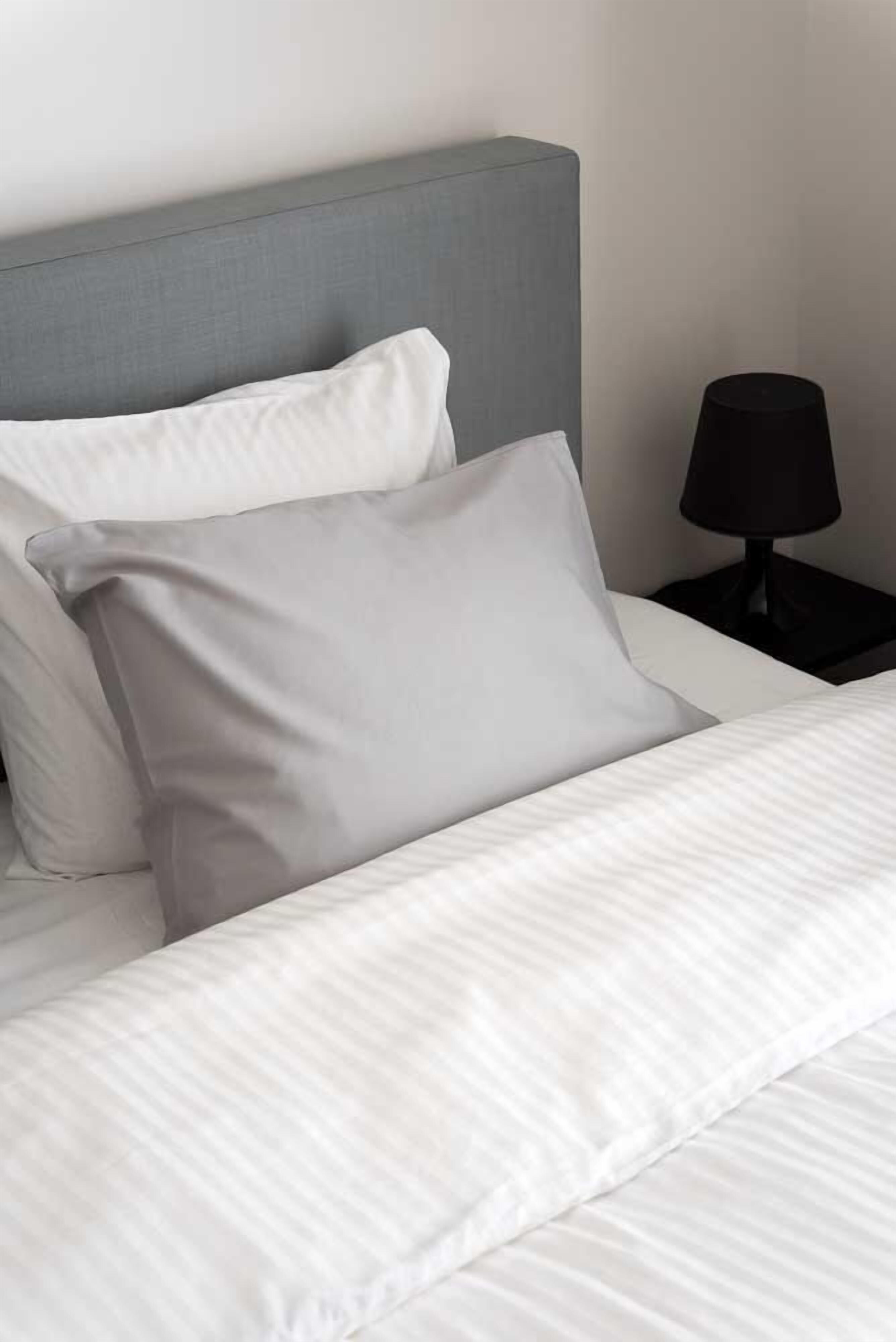 Una cama bien hecha con ropa de cama blanca y dos almohadas, una gris y otra blanca, en una habitación con cabecero gris y una mesita de noche negra con una lámpara.
