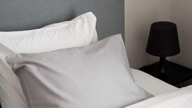 Una cama bien hecha con ropa de cama blanca y dos almohadas, una gris y otra blanca, en una habitación con cabecero gris y una mesita de noche negra con una lámpara.