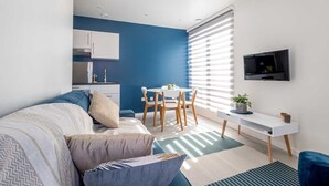 A modern living room with a blue accent wall, a dining area with a white table and chairs, a white TV mounted on the wall, and a kitchen area with white cabinets. - Studio Brussels Airport (Zaventem)