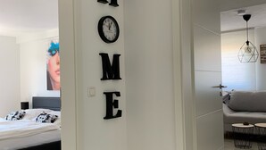 A modern interior with a white wall displaying the word "HOME" in large letters, a bed with black and white bedding, and a grey sofa in the adjacent room. - Modern apartment with prime, kitchen & WLAN (Rudolstadt)