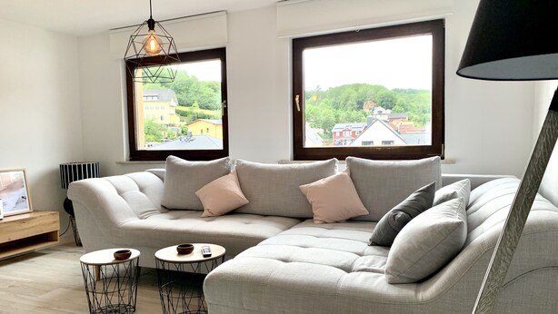A modern living room with a grey sofa, two round side tables, and a large window offering a view of buildings and greenery. - Modern apartment with prime, kitchen & WLAN (Rudolstadt)
