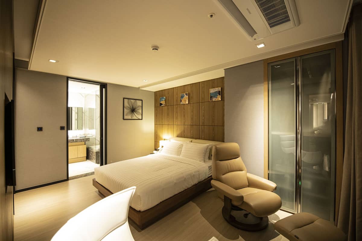 Une chambre d’hôtel moderne dotée d’un grand lit, d’un coin salon avec un fauteuil, d’une douche en verre et d’un mur en panneaux de bois orné d’œuvres d’art.