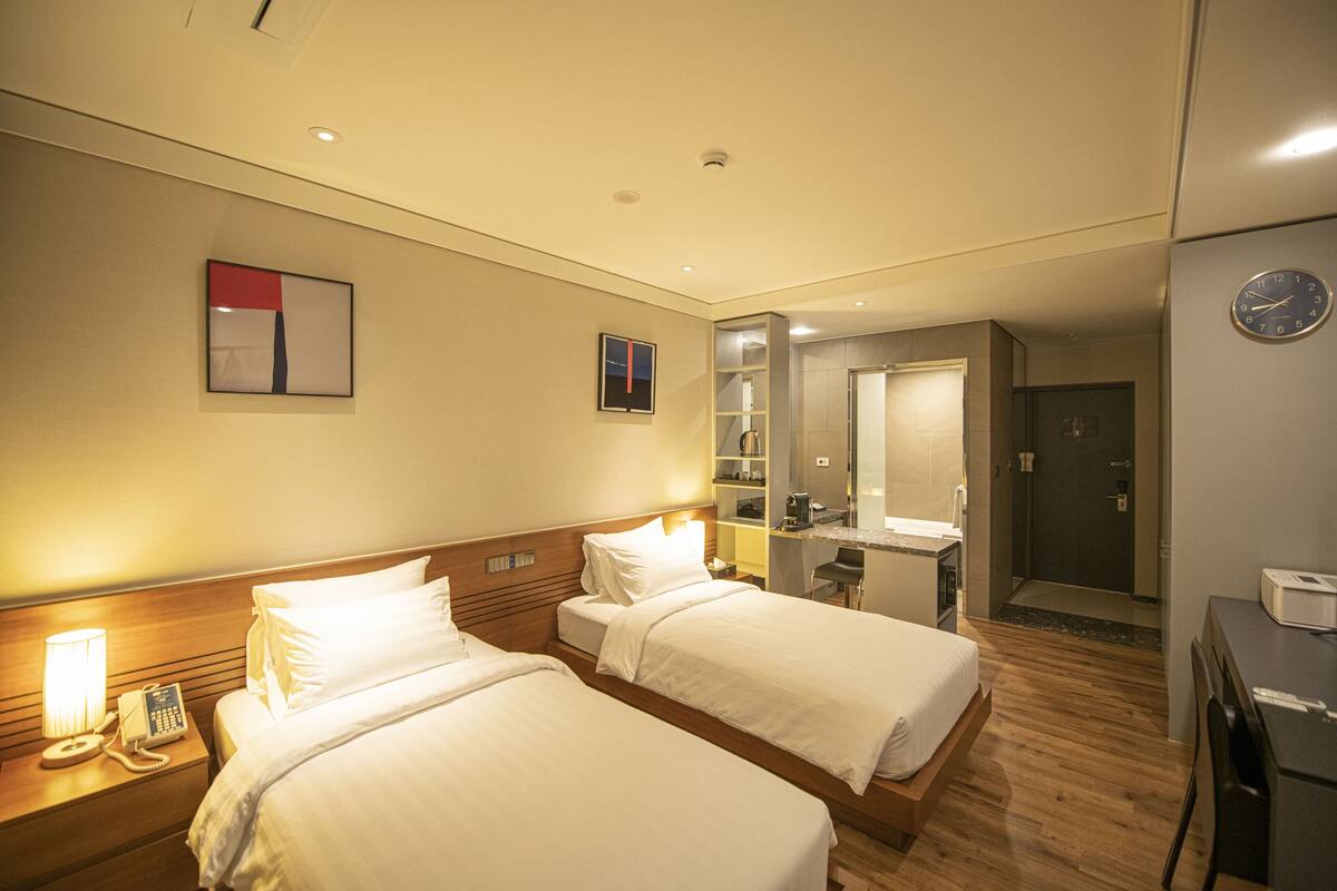 Une chambre d’hôtel avec deux lits, une table de chevet avec une lampe et un téléphone, un petit bureau avec une chaise, un miroir et une porte donnant sur une salle de bain.