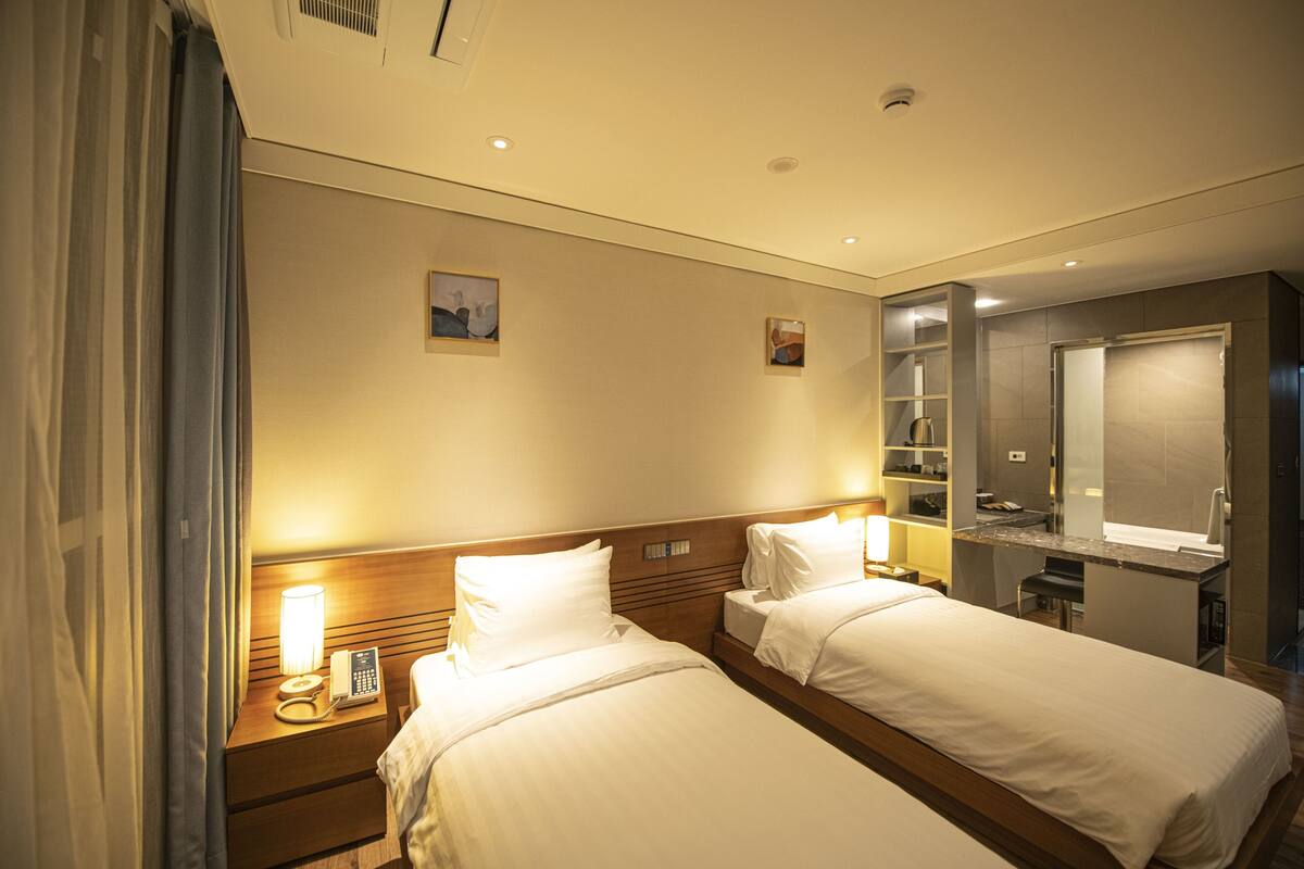 Une chambre d’hôtel avec deux lits, une table de chevet avec une lampe, un miroir et une petite étagère avec quelques objets.