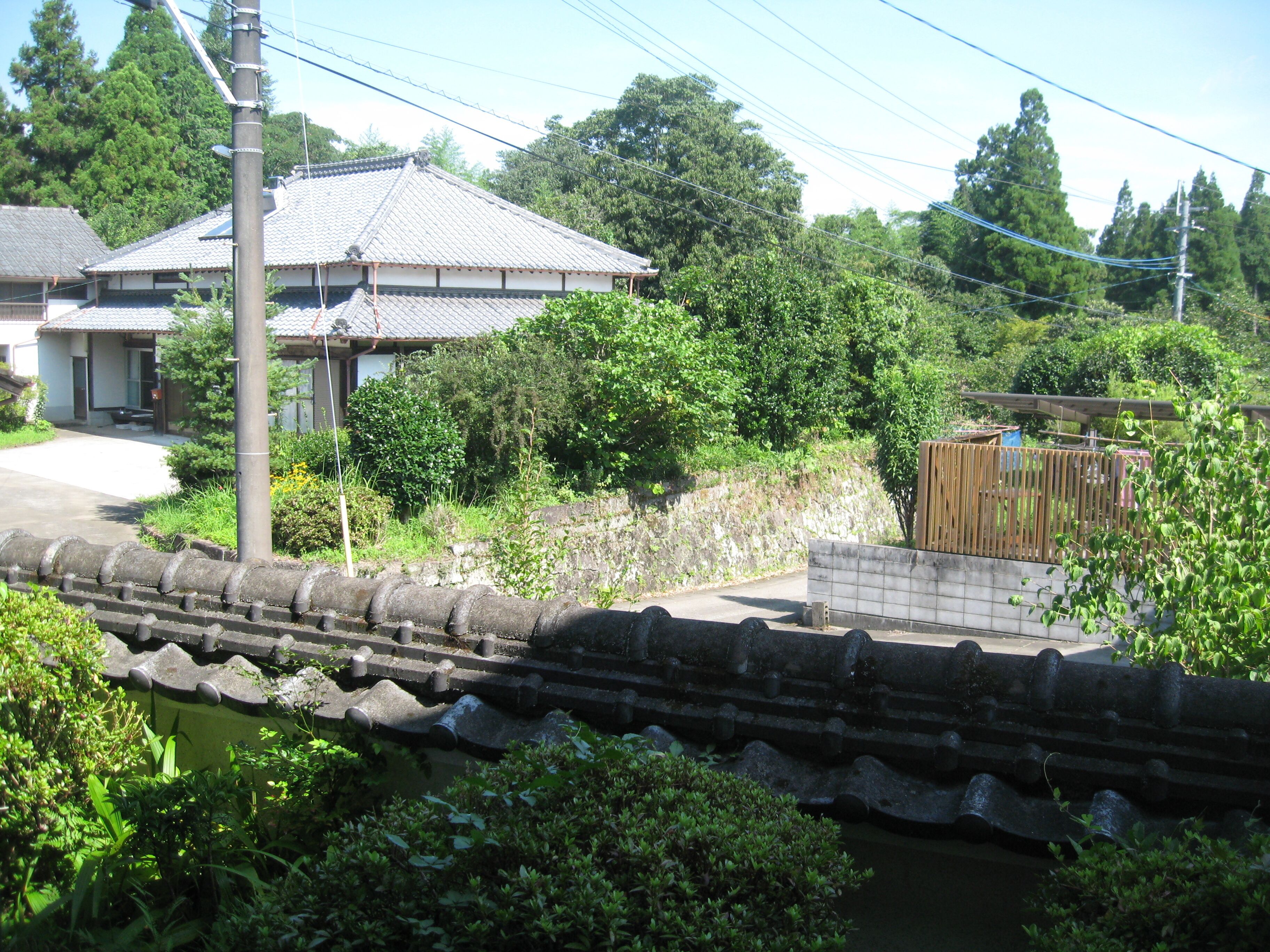 A traditional Japanese house with a tiled roof, surrounded by lush greenery and a stone wall.