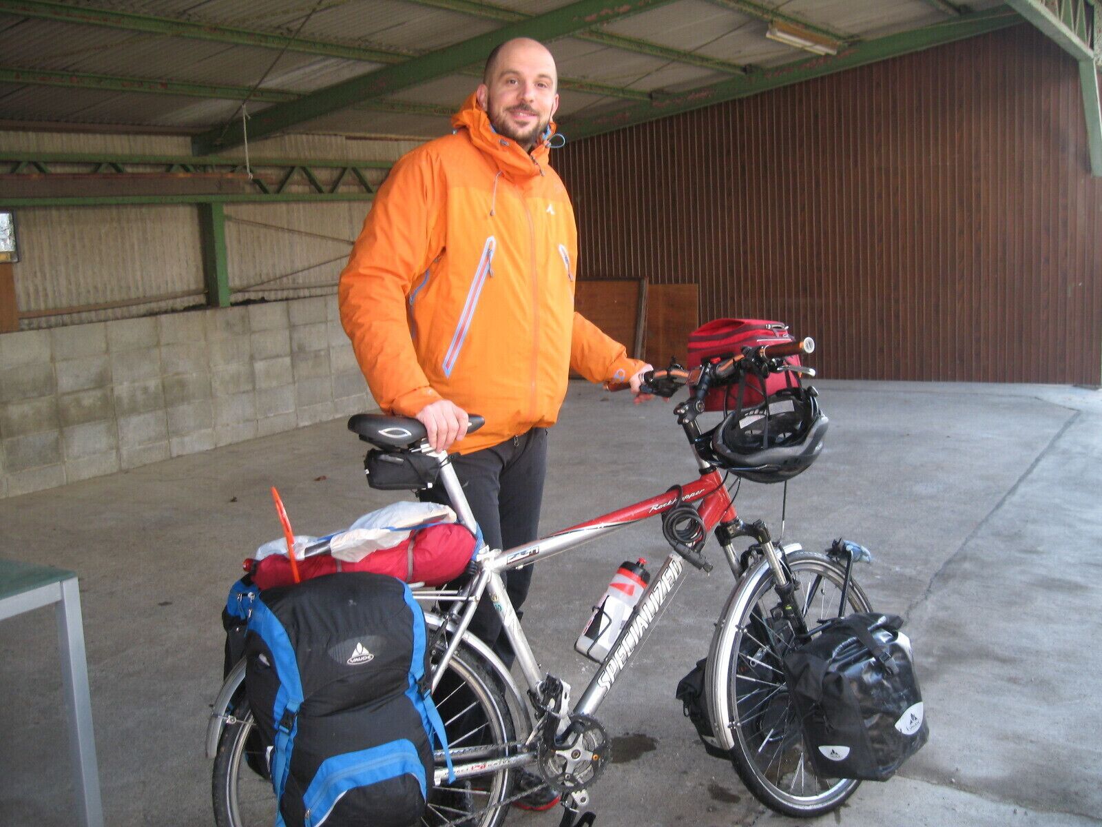 A person standing next to a bicycle with a red frame and a black helmet, wearing an orange jacket.