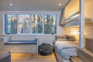 A bedroom with a bunk bed, a window with a mountain view, a bench, and a round pouf.