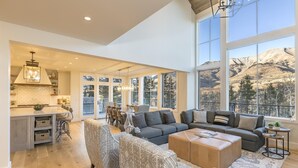 A spacious living area with a large window offering a mountain view, a cozy seating area with patterned chairs, and a kitchen with a modern design. - OVERLOOK HAUS- Mountain Oasis Overlooking San Sophia’s Ridge, Fire Pit & Hot Tub (Mountain Village)