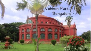 A red colonial-style building with arched windows and a central dome, surrounded by lush greenery and palm trees. - YN Hotels (Bengaluru)