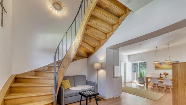 Un interior moderno con una escalera de madera, un sofá gris, una mesa de ajedrez y un comedor con sillas.