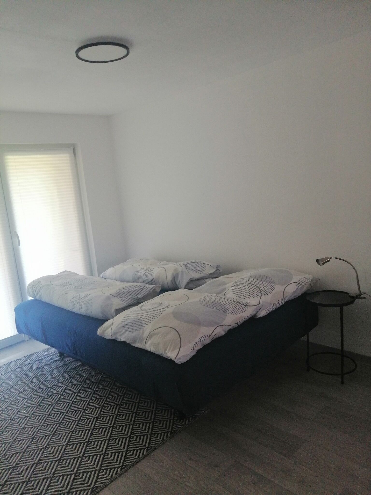 Quarto com cama, mesinha de cabeceira, abajur e tapete estampado.