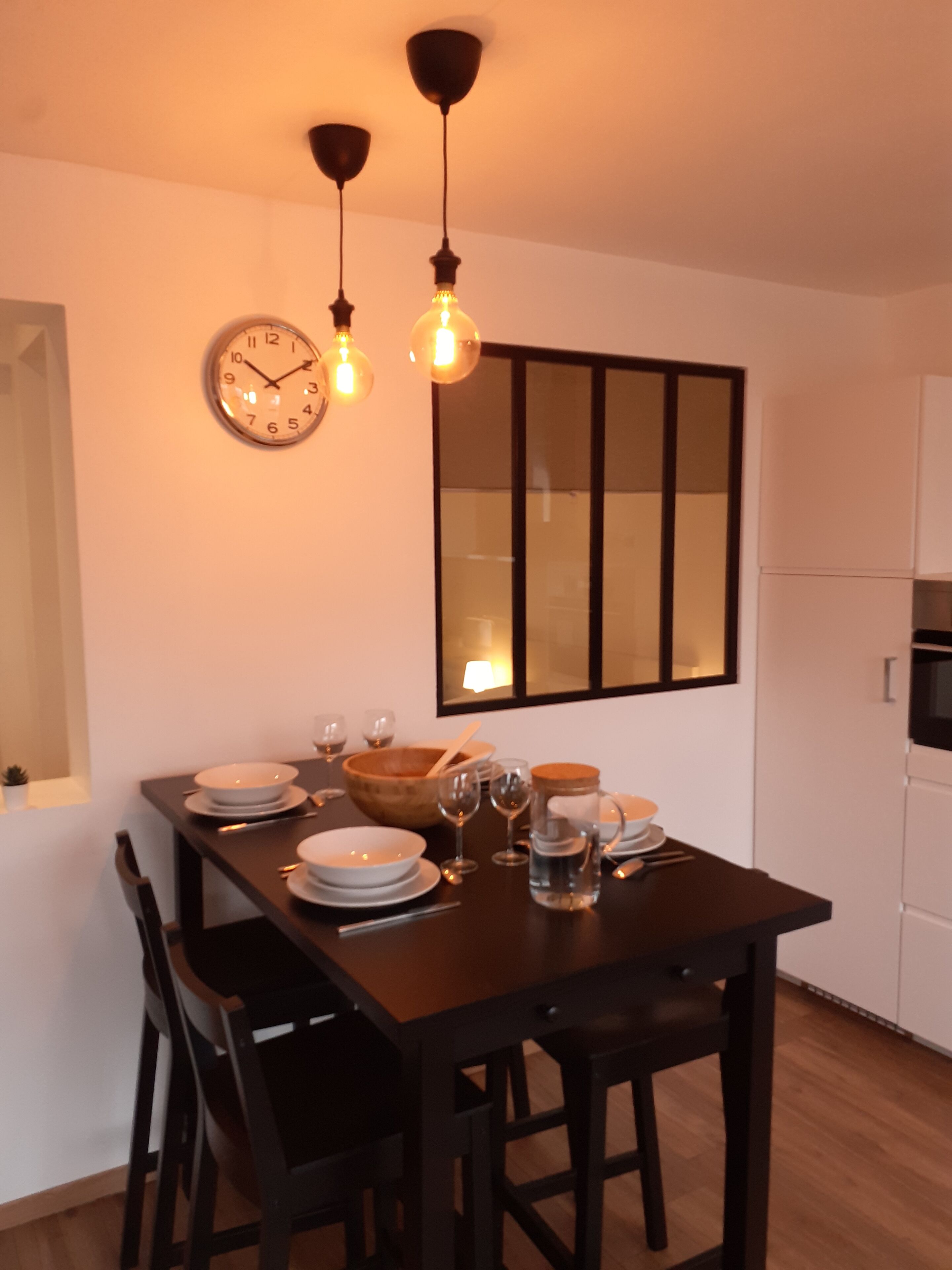 A dining area with a dark wooden table set for four, surrounded by chairs. A wall clock and pendant lights are visible above the table.