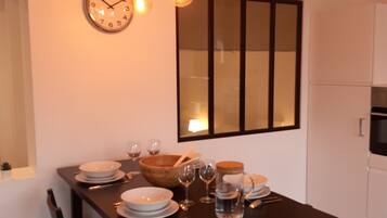 A dining area with a dark wooden table set for four, surrounded by chairs. A wall clock and pendant lights are visible above the table.