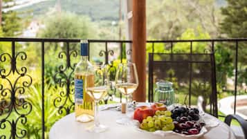 A table with a bottle of white wine, two wine glasses, a plate of fruit, and a glass jar on a balcony with a metal railing and greenery in the background.