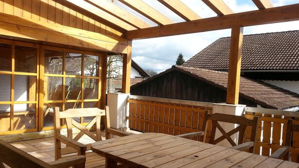 Une terrasse en bois avec une table et des bancs, un espace couvert avec des fenêtres, et une vue sur une maison avec un toit en tuiles.