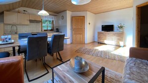 Een ruime woonkamer met een houten plafond, een eettafel met stoelen, een televisie op een houten kast, en een keuken op de achtergrond.