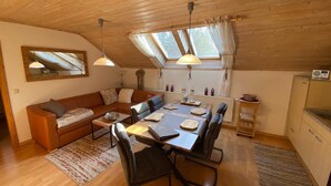 Een gezellige, goed verlichte eetruimte met een houten tafel, stoelen en een bank. Er zijn plafondlampen, een raam met gordijnen en een spiegel aan de muur.