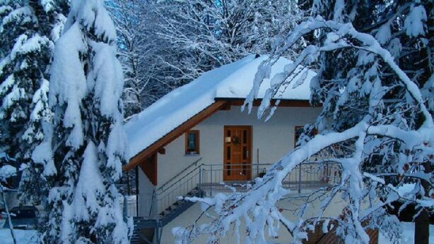 Een huis bedekt met sneeuw, met een houten trap naar een voordeur, omringd door sneeuwzware bomen.