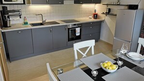Una cocina moderna con armarios grises, un refrigerador de acero inoxidable, un microondas y un horno. Hay una mesa de comedor con un tazón de fruta y una copa de vino.