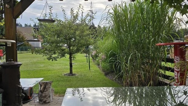 Un porche cubierto con una mesa, una lámpara colgante y vistas de un jardín con césped, árboles y una estructura roja.