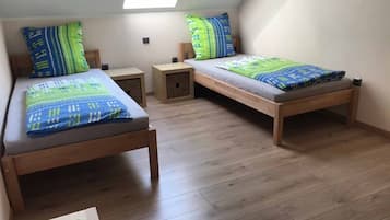 A dormitory room with two wooden beds, each with a matching headboard and footboard. There are two bedside tables, one on each bed, and a small storage box under the headboard of the bed on the right. The room has a sloped ceiling with skylight windows, allowing natural light to enter.
