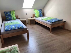 A dormitory room with two wooden beds, each with a matching headboard and footboard. There are two bedside tables, one on each bed, and a small storage box under the headboard of the bed on the right. The room has a sloped ceiling with skylight windows, allowing natural light to enter. - Ferienwohnung Anna (Moosbach)