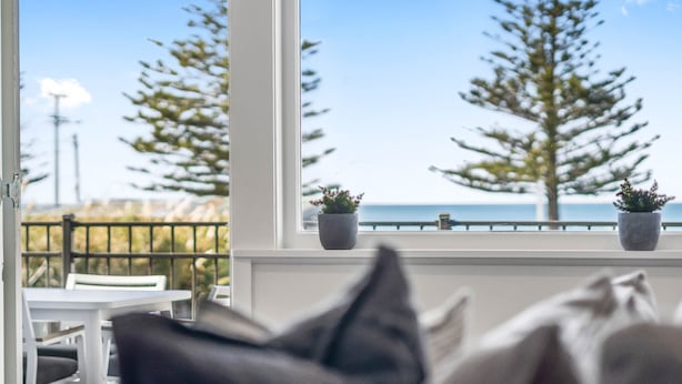 Un balcon avec vue sur la mer et les arbres, deux plantes en pot et une table avec des chaises.