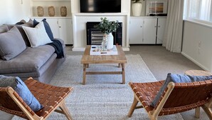 Un salon avec un canapé gris, une table basse en bois, deux fauteuils en osier marron, un téléviseur à écran plat placé au-dessus d’une cheminée et une plante en pot.