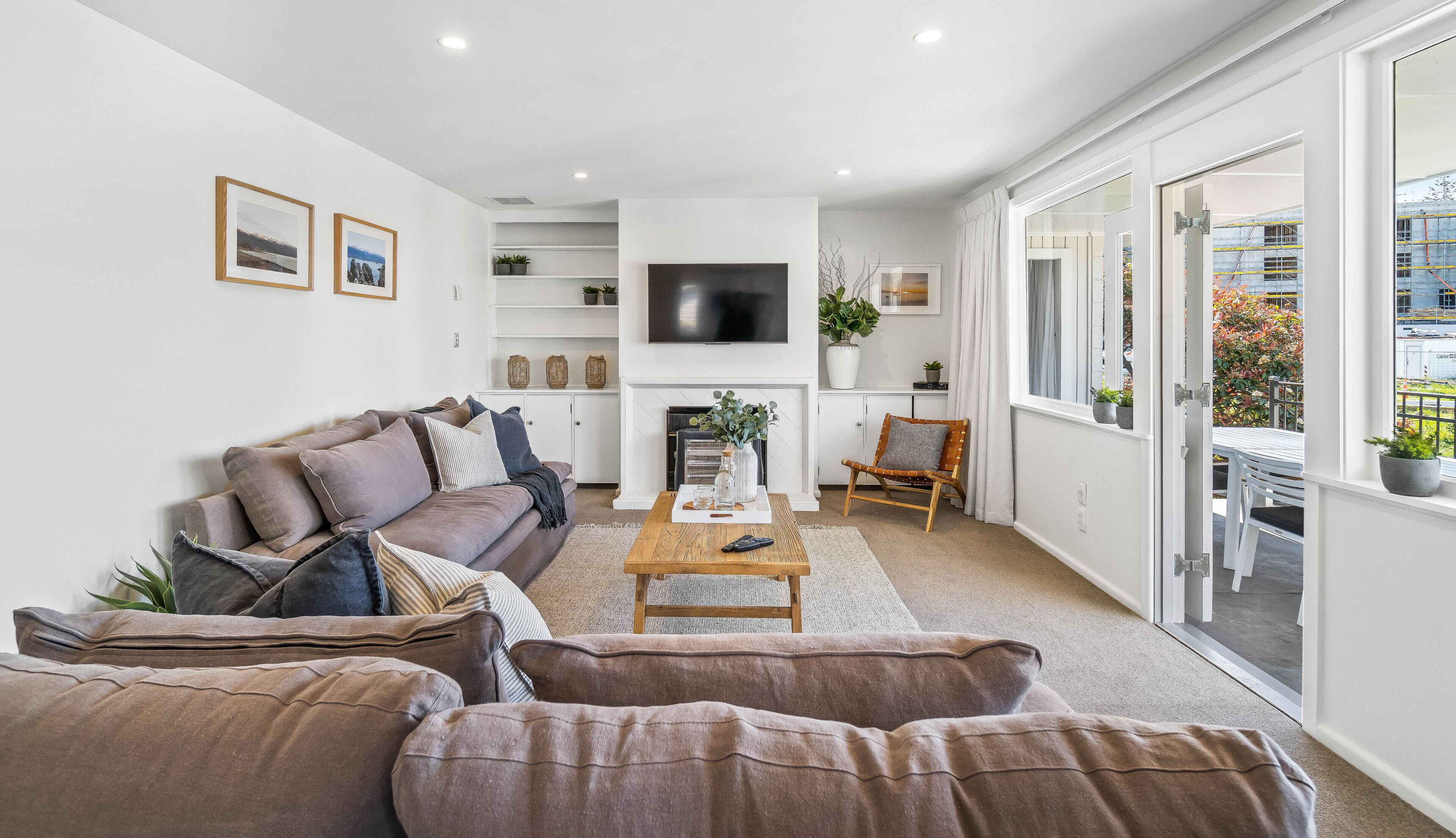 A modern living room with a sofa, coffee table, and a fireplace. There is a TV mounted on the wall and a window with a view of the outside.