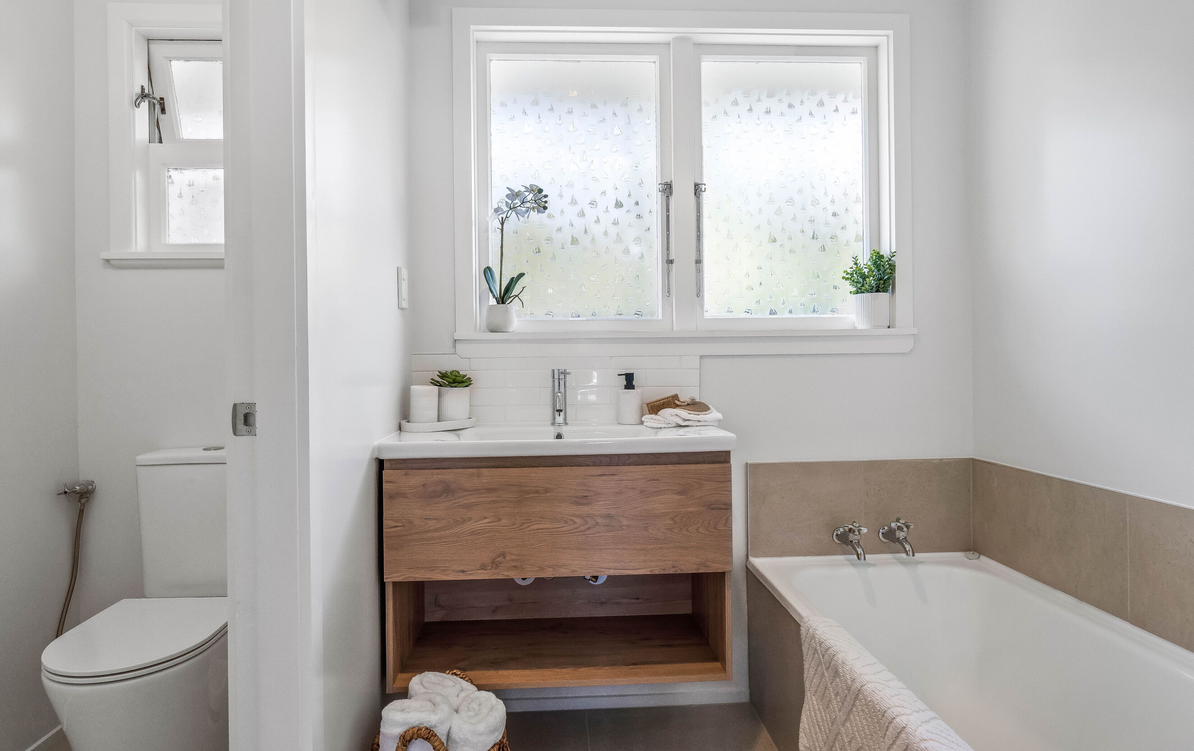 A modern bathroom with a white toilet, a wooden vanity, a bathtub, and a window with frosted glass.