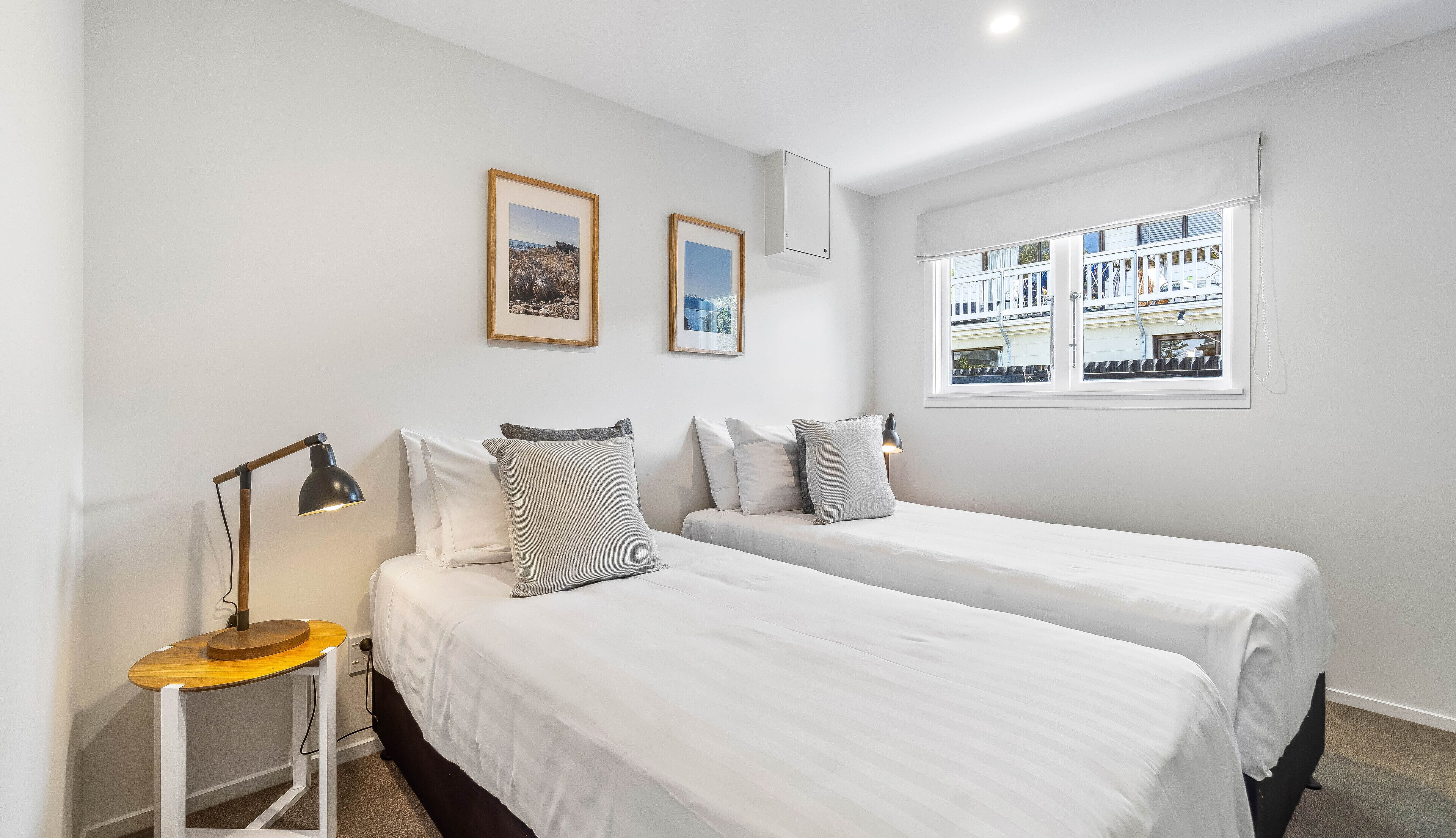 A bedroom with two beds, a bedside table with a lamp, and a window with a view of buildings.