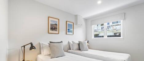 A bedroom with two beds, a bedside table with a lamp, and a window with a view of buildings.