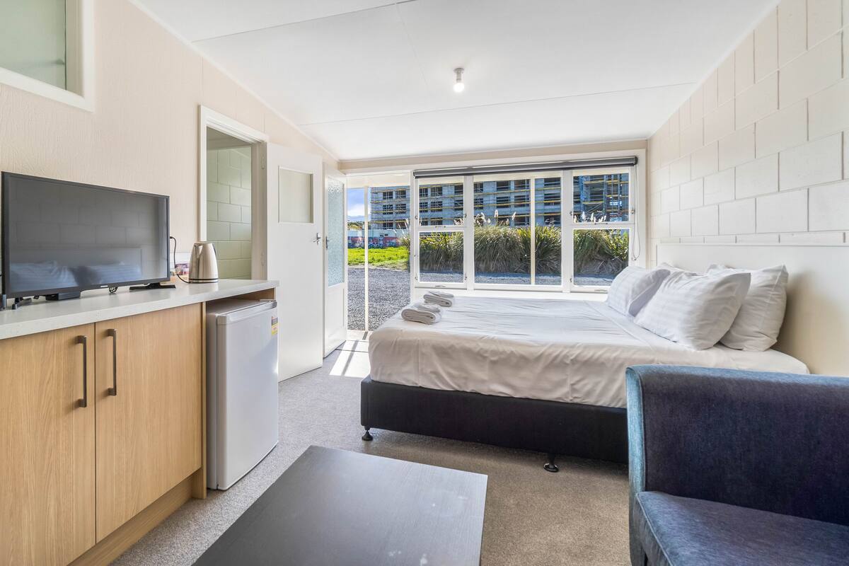 Une chambre d’hôtel compacte comprenant un coin cuisine, un lit, un espace salon et une grande fenêtre donnant sur l’extérieur.