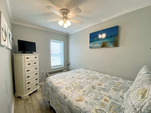 A bedroom with a bed, a dresser, a ceiling fan, a window, and a painting on the wall. - Beachview 216 (Gulf Shores)