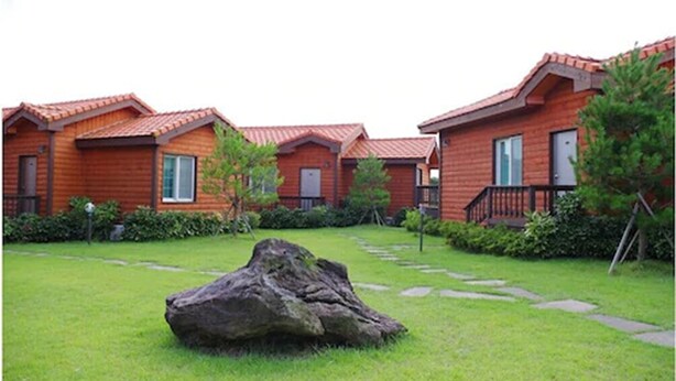 A row of log cabin-style cabins with red roofs and wooden exteriors, surrounded by well-maintained lawns and a large rock in the foreground. - Haemaji Pension (Seogwipo)