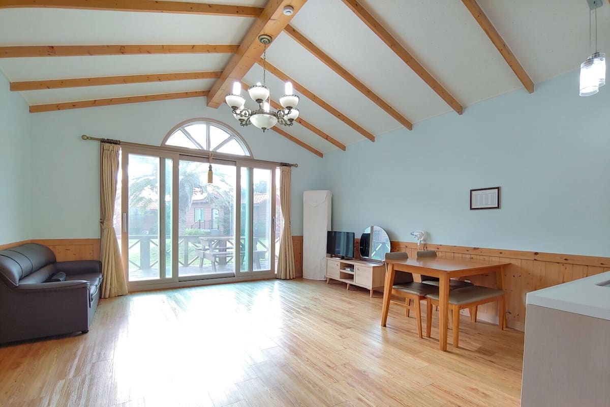 Una habitación amplia con techo de madera, un gran ventanal corredero que da a un espacio exterior, un sofá, una mesa de comedor con sillas, un televisor y un mueble auxiliar.