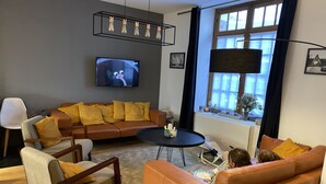 A modern living room with a brown leather sofa, a grey armchair, and a black coffee table. A flat-screen TV is mounted on the wall, and there are two framed pictures on the wall.