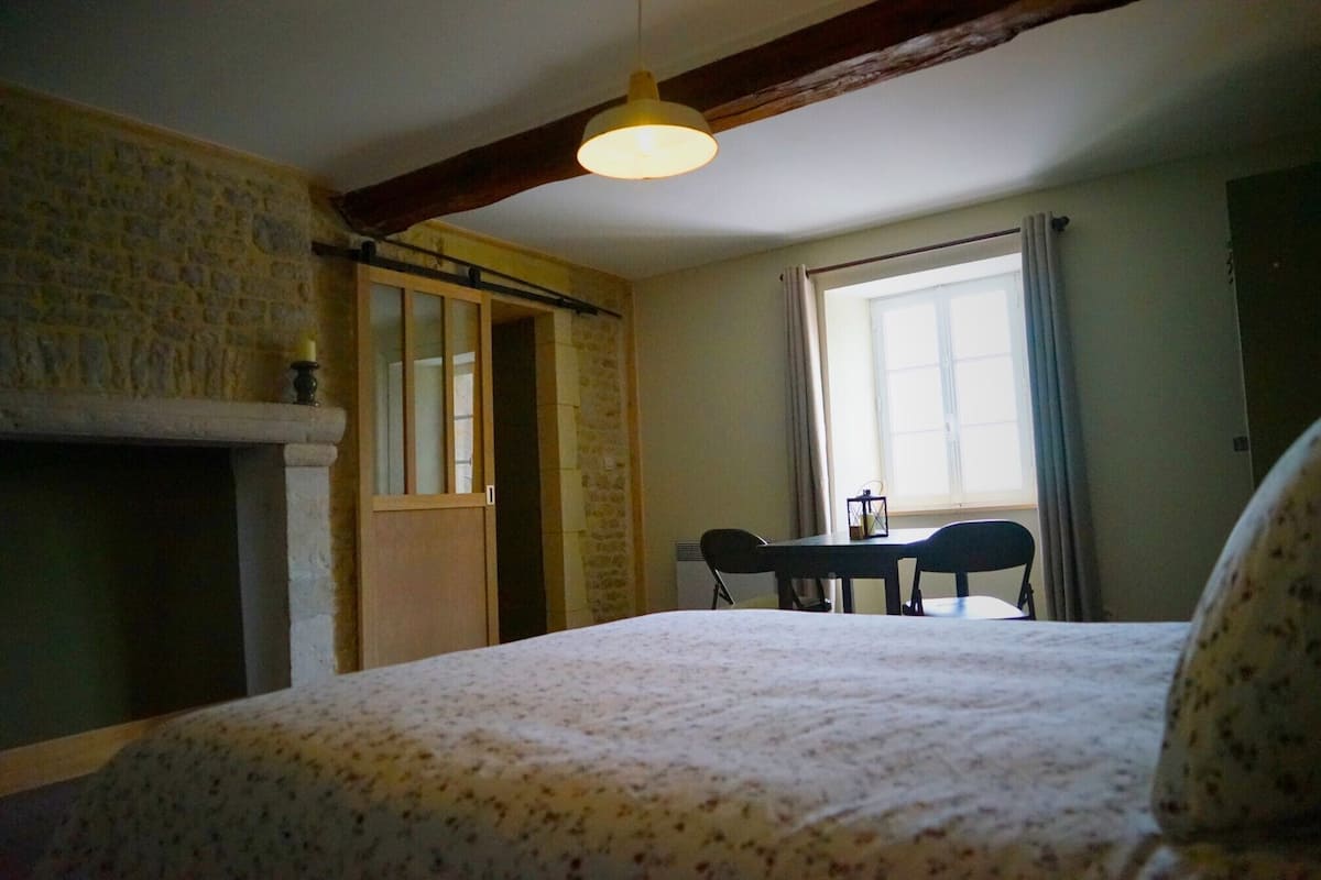 Een kamer met een bed, een tafel met stoelen, een raam met gordijnen en een stenen muur.