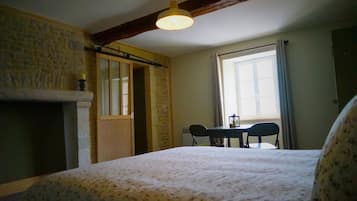 A room with a bed, a table with chairs, a window with curtains, and a stone wall.