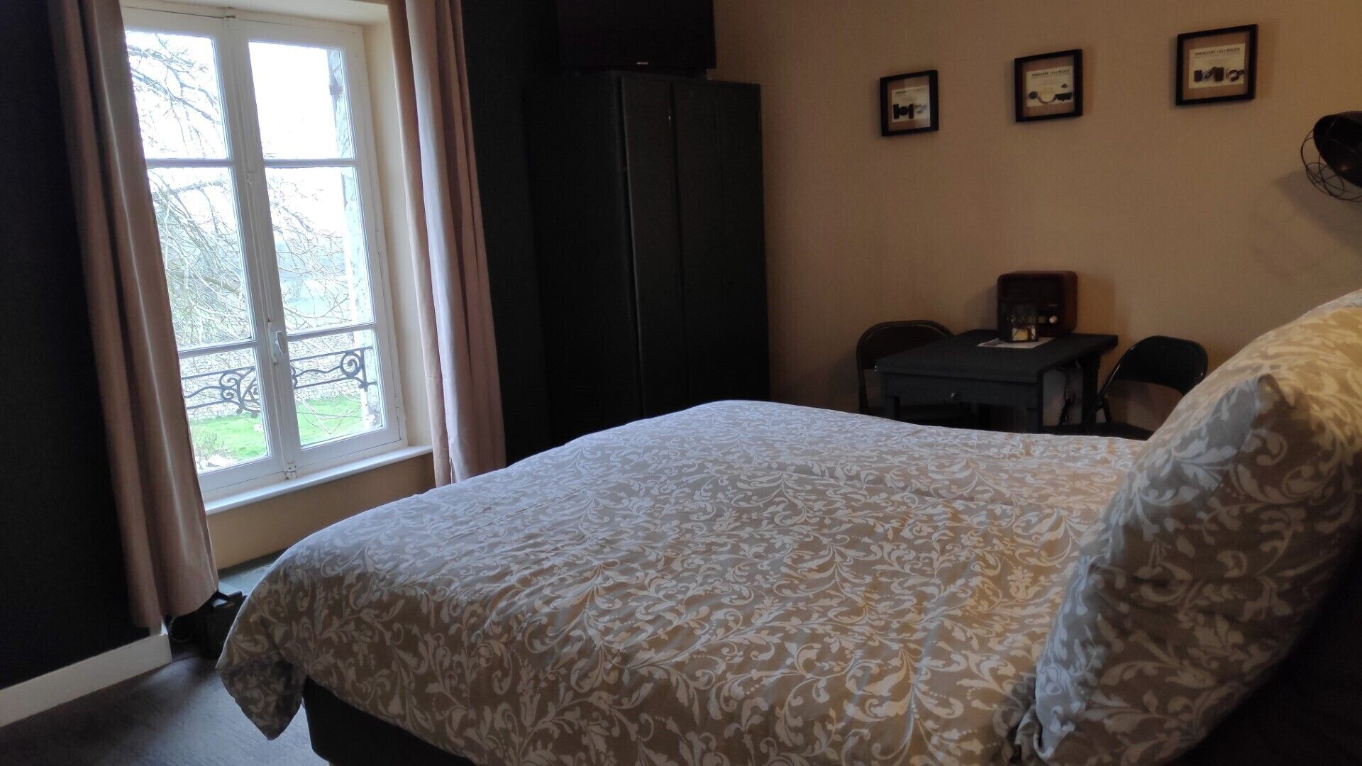 Een slaapkamer met een bed, een raam met gordijnen, een donkere kast, een kleine tafel met een lamp en drie ingelijste foto’s aan de muur.