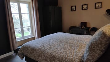 Een slaapkamer met een bed, een raam met gordijnen, een donkere kast, een kleine tafel met een lamp en drie ingelijste foto’s aan de muur.