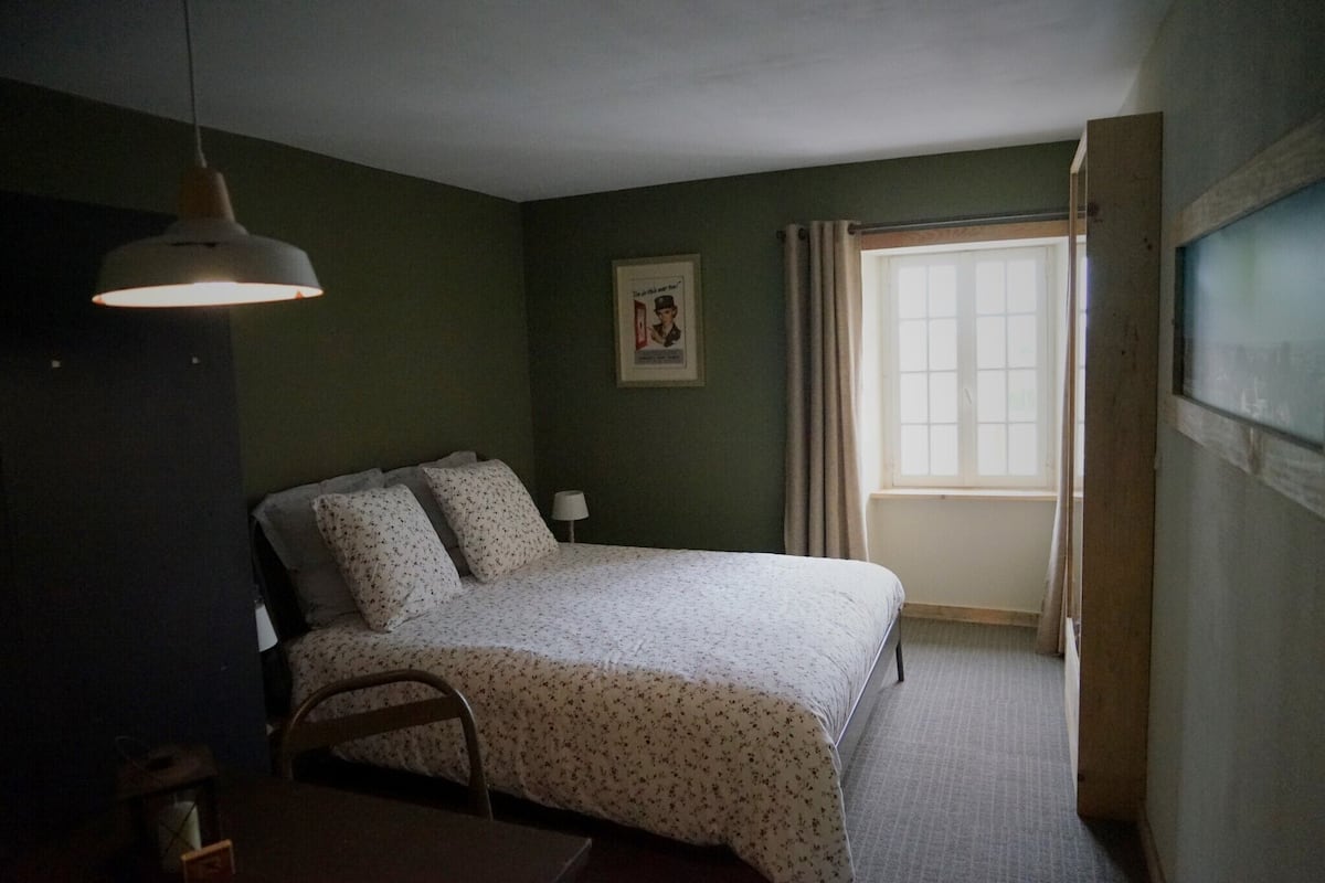 Een slaapkamer met een bed, een hanglamp, een raam met gordijnen, en een lijst met een afbeelding aan de muur.