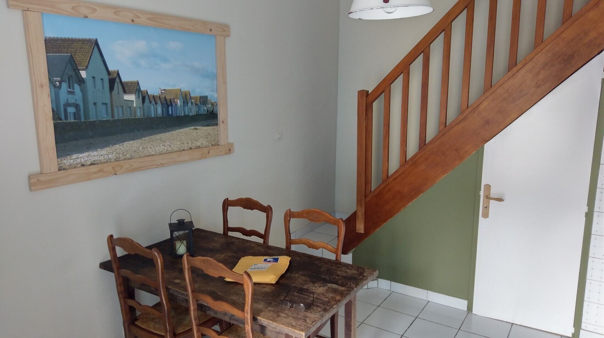 Un comedor con una mesa y sillas de madera, un cuadro enmarco que muestra casas, y una escalera con barandilla de madera.