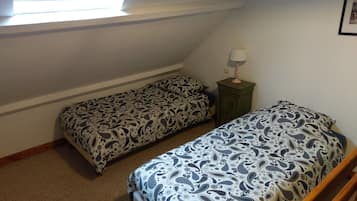 Um quarto pequeno e simples, com duas camas de solteiro, uma mesa de cabeceira com abajur e uma janela que oferece iluminação natural.